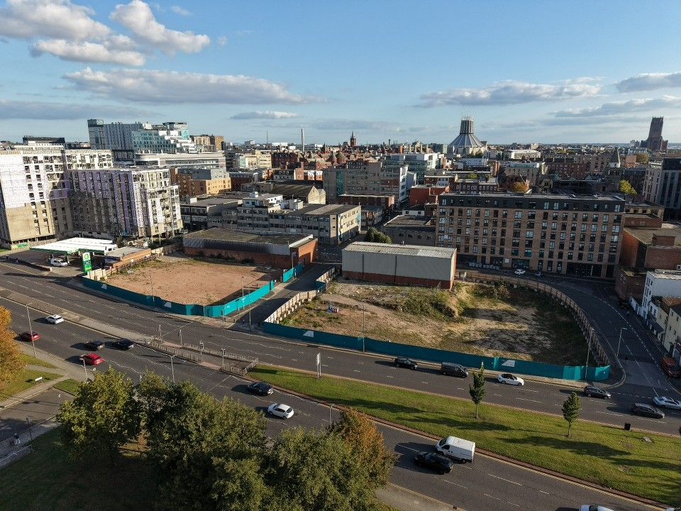 Aerial photograph of a development site in Liverpool, showing cleared land beside a main road with the city skyline and Liverpool Metropolitan Cathedral in the background.